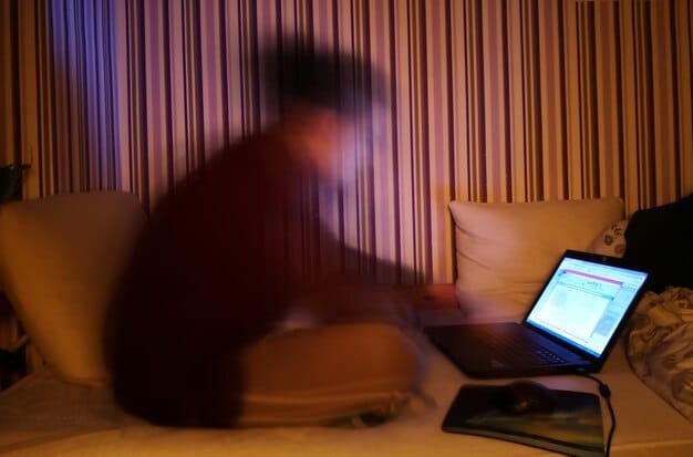 A person in a dimly lit room typing on a computer, emphasizing secrecy and anonymity while reporting a crime.